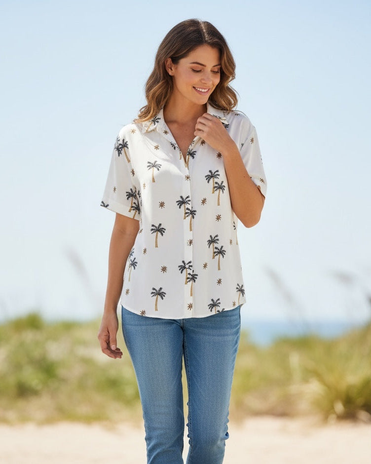 Woman wearing a white blouse with palm tree pattern and blue jeans on a beach.